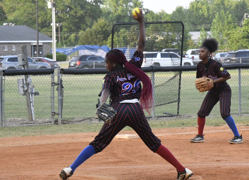 A softball pitcher winds up to throw the ball, while an infielder is ready, both wearing black and red uniforms on a dirt field.