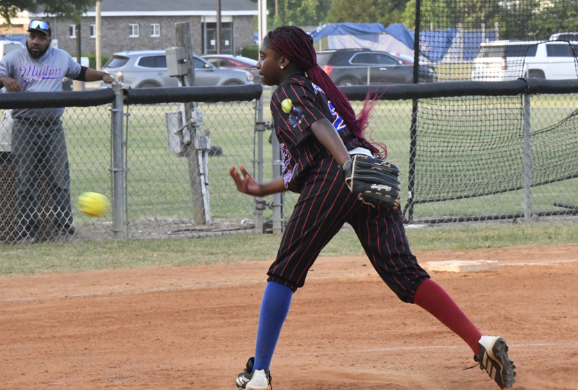 A softball pitcher in a black uniform with red stripes throws a pitch on a field; spectators and cars are visible in the background.