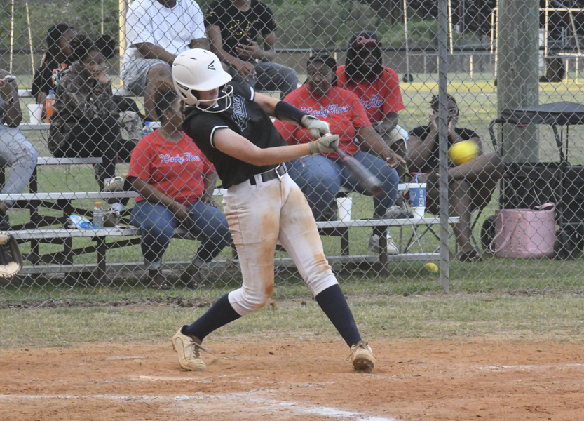 A softball player in a helmet swings at a pitch during a game, with spectators watching from the bleachers behind a chain-link fence.