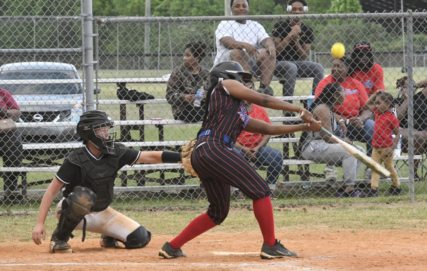 A softball player swings at a pitch, with a catcher in position and spectators watching from the bleachers behind a chain-link fence.