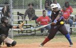 Softball player in black and red uniform swings at a pitch while the catcher prepares to catch the ball, with spectators watching behind a chain-link fence.