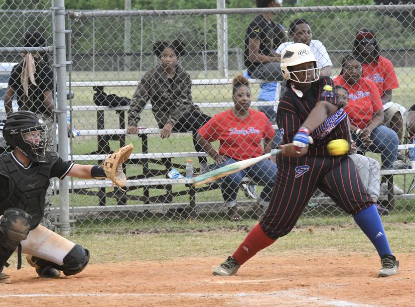 Softball player in black and red uniform swings at a pitch while the catcher prepares to catch the ball, with spectators watching behind a chain-link fence.
