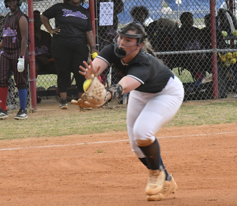 Softball player in action fielding a ground ball, wearing a helmet and glove, with team members and spectators in the background near a chain-link fence.