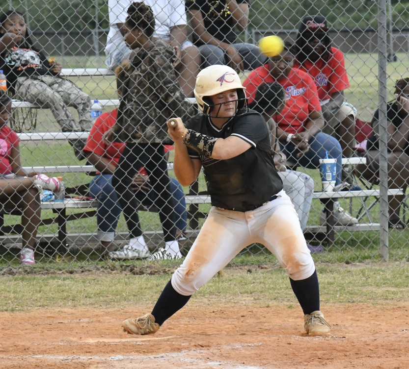 A softball player in batting stance, wearing a helmet, prepares to swing at an incoming pitch, with spectators seated behind a chain-link fence watching the game.