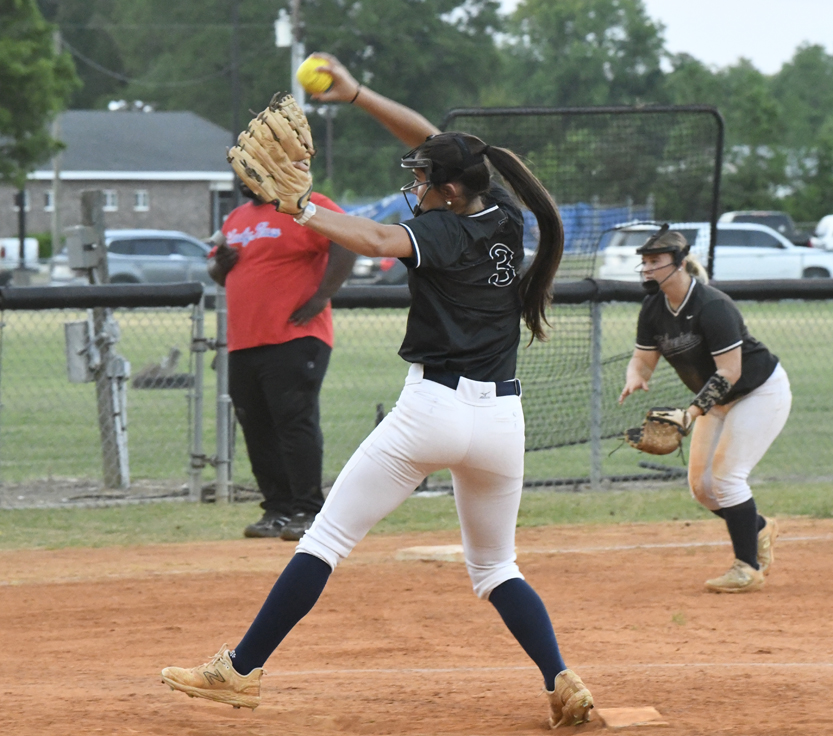 A softball pitcher in mid-throw on a dirt infield, with a teammate and coach in the background, during a daytime game.