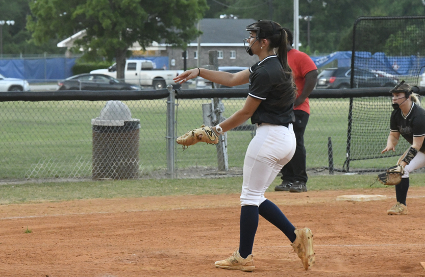 Softball pitcher in mid-throw on a dirt field, wearing protective face mask, with another player and a coach in the background.