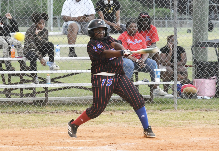 A softball player in a black and red uniform swinging a bat, with spectators sitting on bleachers behind a chain-link fence in the background.
