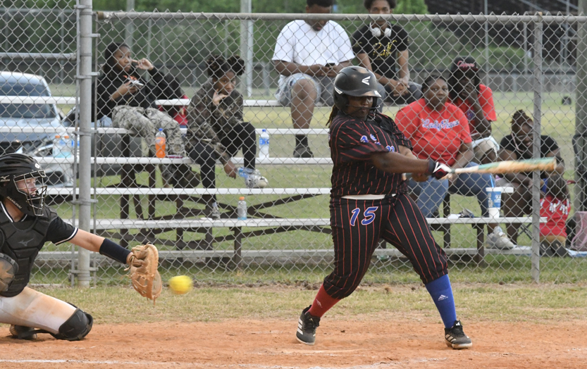 Softball player swinging bat at a yellow ball, with catcher in gear and spectators sitting behind a chain-link fence on bleachers.