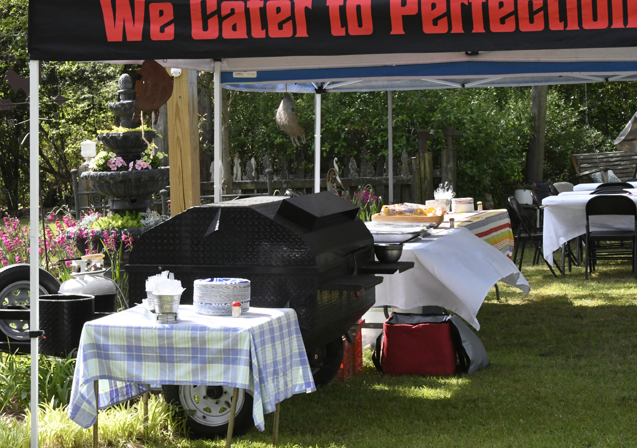Outdoor catering setup with covered tables, a large black barbecue smoker, and a sign that reads "We Cater to Perfection" under a canopy in a grassy area.