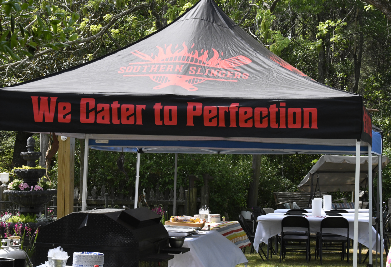 Outdoor catering tent with "We Cater to Perfection" printed on the canopy. Tables with food and drinks are set up underneath in a park-like setting.