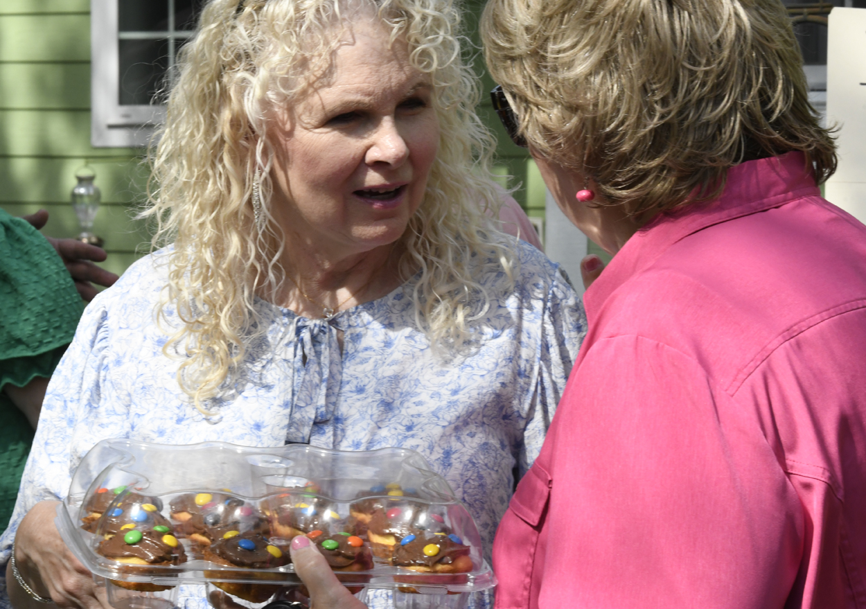 Two women talking outdoors, one with curly blonde hair holding a tray of cookies wrapped in plastic, both appear to be enjoying a friendly conversation.