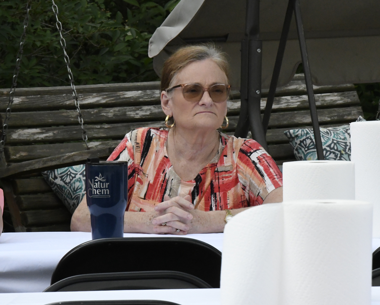 Woman wearing sunglasses and a colorful shirt sits at an outdoor table with a blue tumbler and rolls of paper towels nearby.