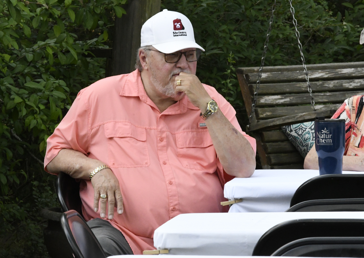 older man wearing a pink shirt and white cap sits outdoors at a table, appearing to be deep in thought.
