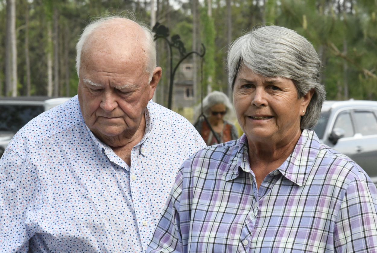 older man and woman stand outdoors, both wearing casual shirts. The woman is smiling while the man looks down. Trees and another person are visible in the background.