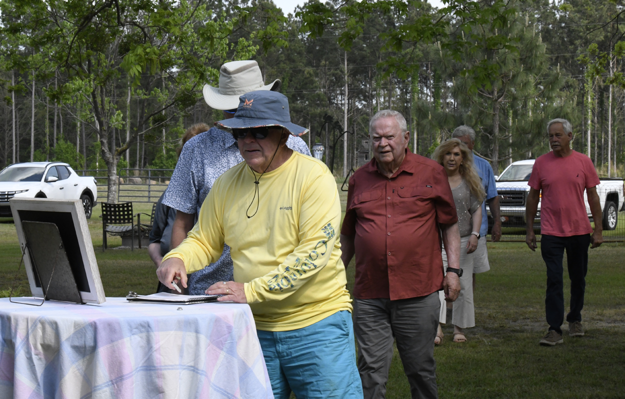 A group of people standing outdoors, one man in a yellow shirt and sun hat writing on paper atop a table, trees in the background.