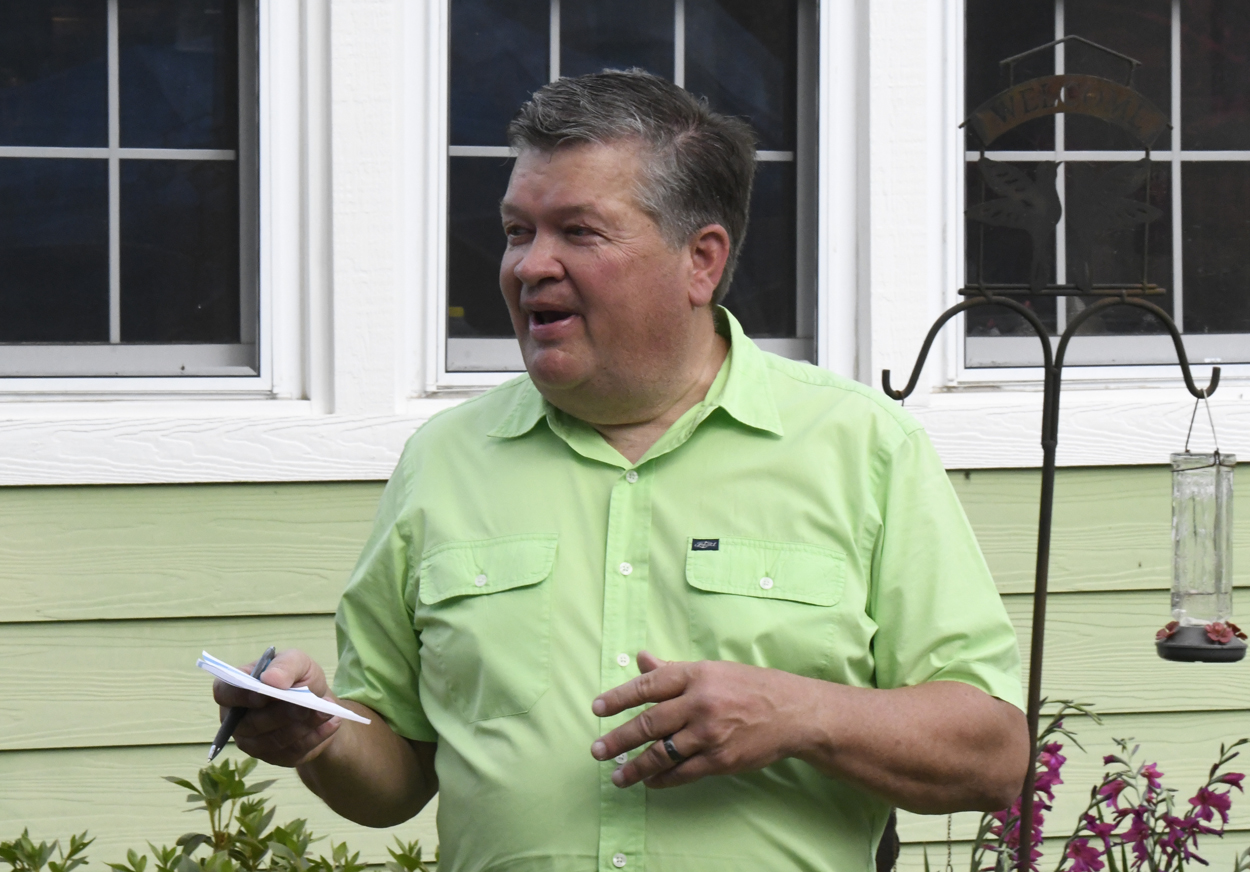 Man in a light green shirt stands outdoors, holding a notepad and pen, speaking in front of a house with white-framed windows.
