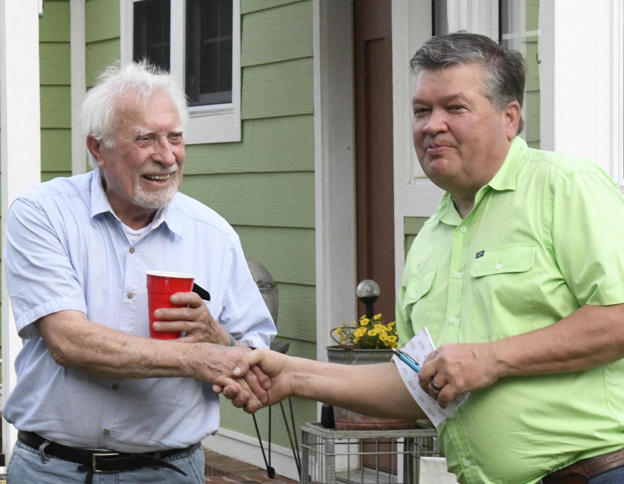 Two men standing outside, shaking hands and smiling, with a green house in the background. One man holds a red cup.