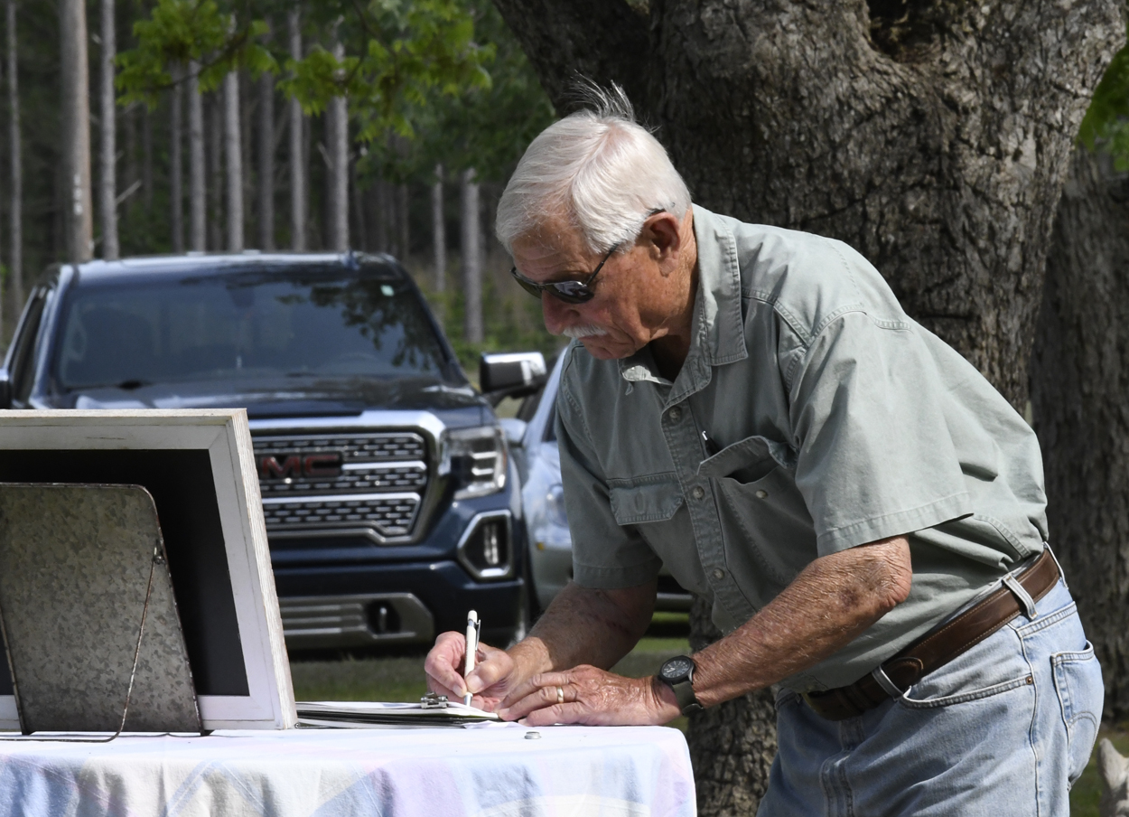 Older man wearing sunglasses writes on a piece of paper at an outdoor table, with parked cars and trees visible in the background.