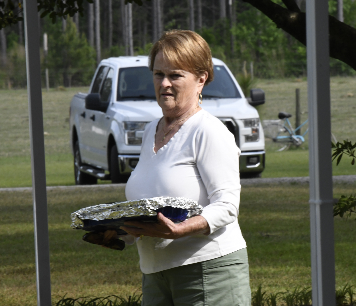 Woman outdoors holding a covered dish, with a white pickup truck and green grass visible in the background.