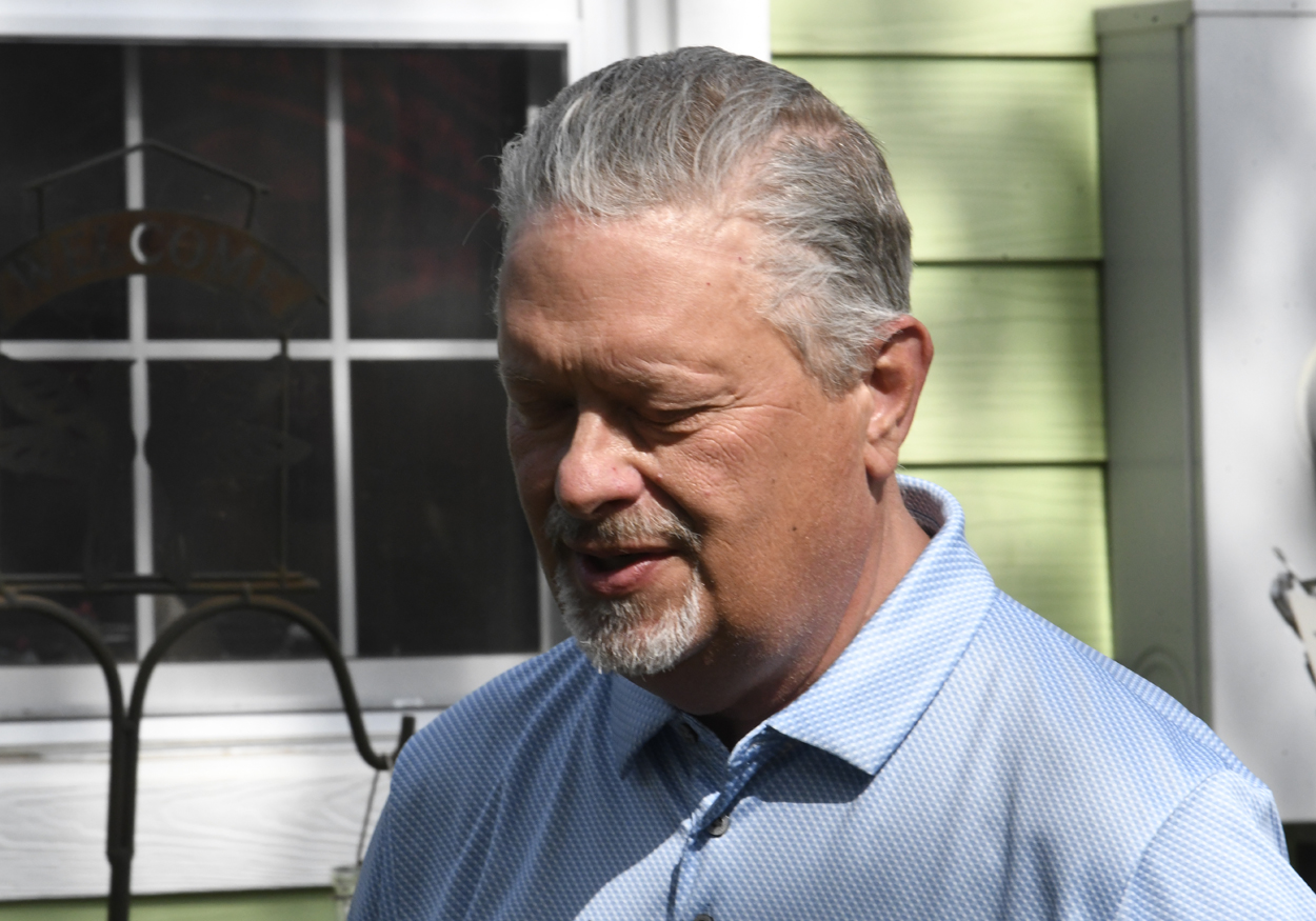 Older man with gray hair and goatee, wearing a light blue collared shirt, standing outside near a window and green wall in sunlight.