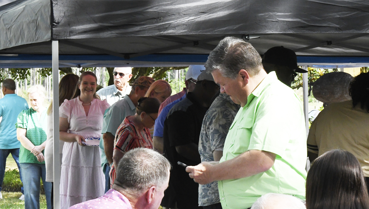 A group of people gathers under a tent outdoors; one man in a green shirt appears to be writing or checking something.