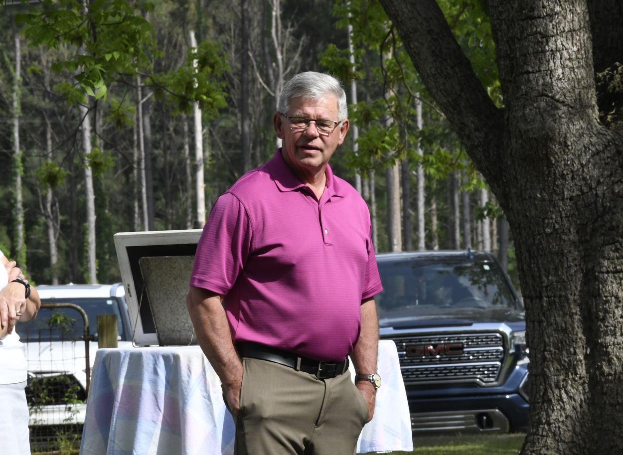 Older man wearing a pink polo shirt and khaki pants standing outdoors near parked vehicles, with a table covered by a white cloth in the background.