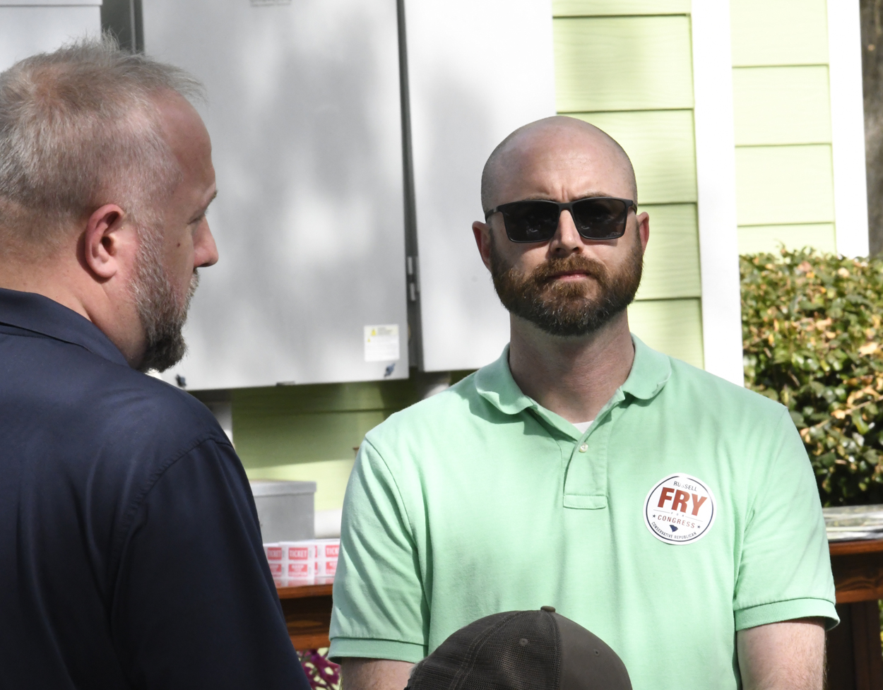 Two men talking outdoors, one wearing sunglasses and a green shirt with a name tag.