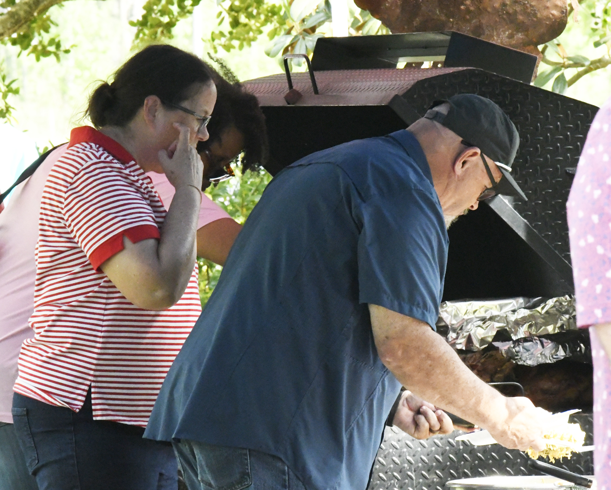 Two people standing outdoors near a barbecue grill; one is cooking while the other observes, both focused on the task under leafy trees.