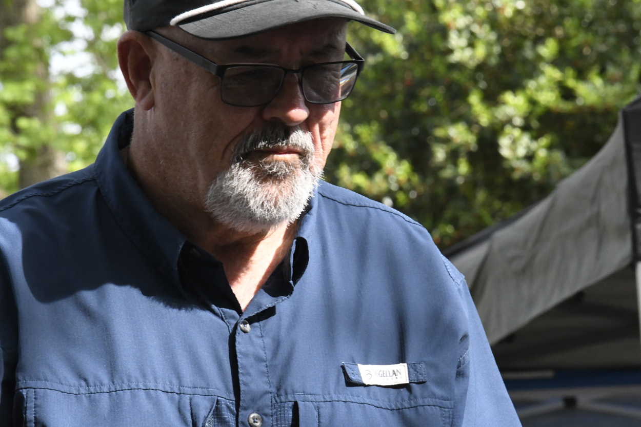 Older man wearing glasses, a hat, and a blue shirt standing outdoors in sunlight, with trees and greenery in the background.