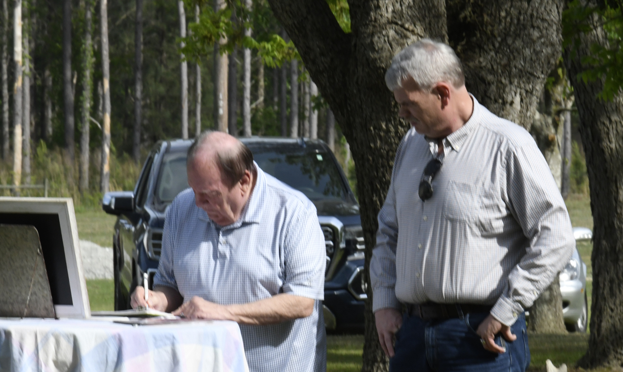 Two older men outdoors, one writing on a table covered with a cloth, the other standing and watching, with trees and cars in the background.