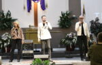 Three people sing on a church stage, standing in front of a cross, with musical equipment and plants visible around them.