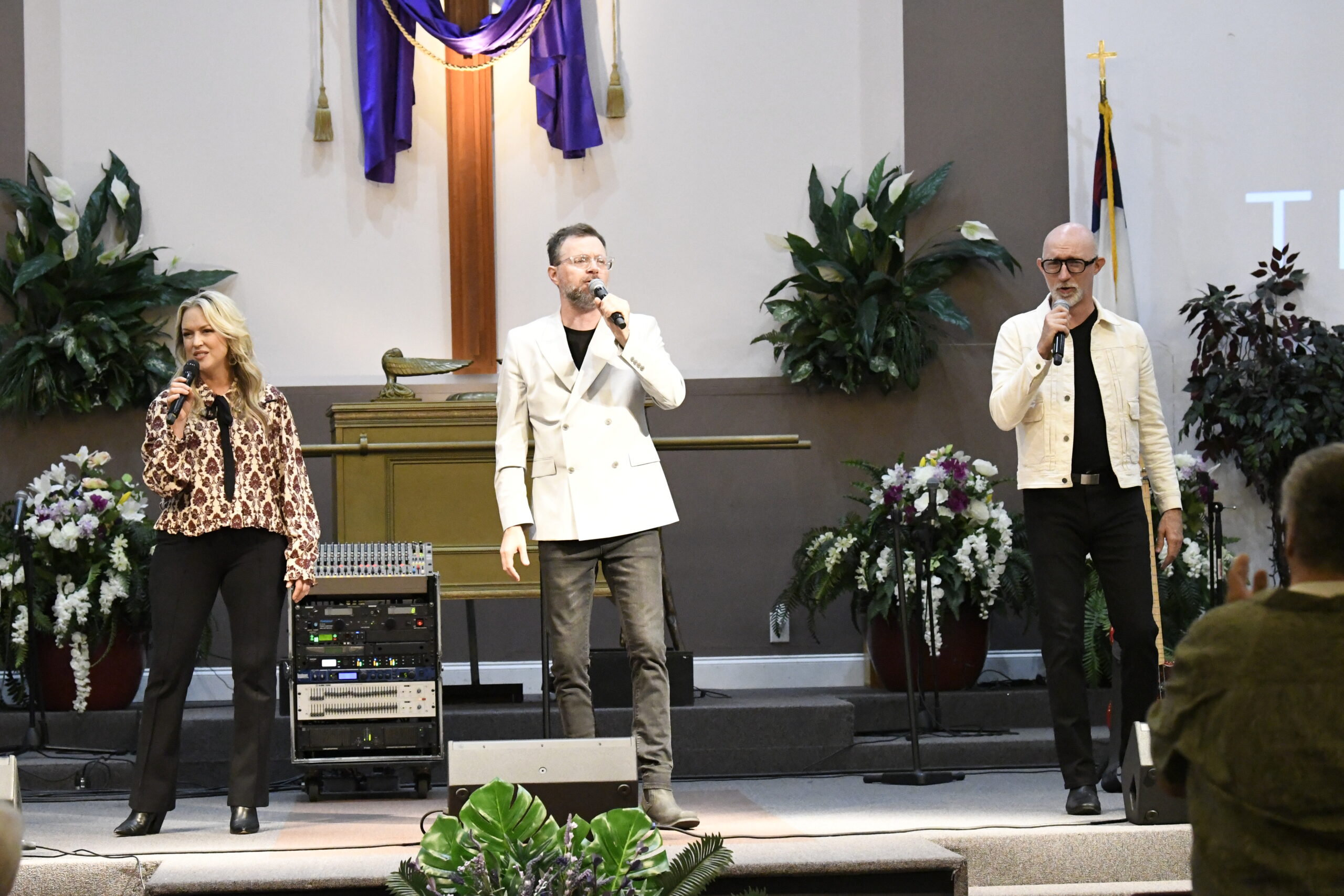 Three people sing on a church stage, standing in front of a cross, with musical equipment and plants visible around them.