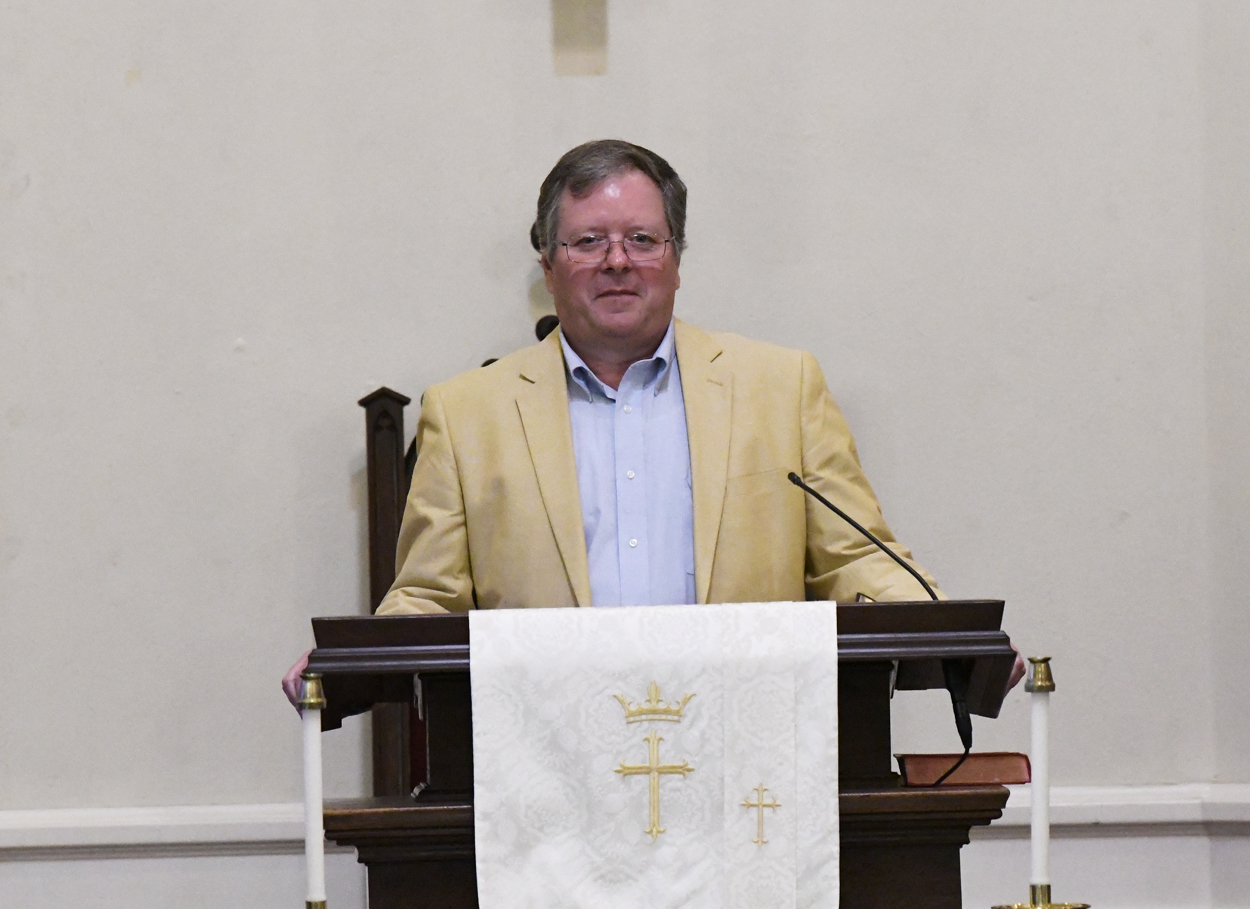 A man in a tan jacket stands at a church pulpit with a white cloth displaying gold religious symbols.
