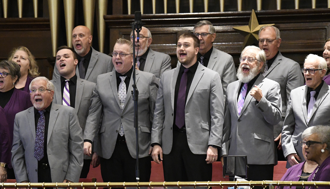 A group of men in gray suits sing together in a choir on stage, standing in front of a microphone and a wooden background.
