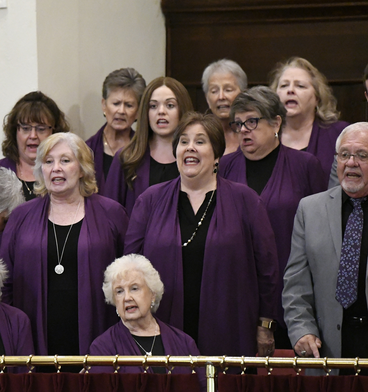 A choir of older adults sings together, most wearing matching purple attire, standing in rows inside a building.