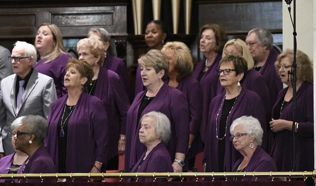A choir of women in matching purple outfits sings together in a church setting, with organ pipes visible in the background.
