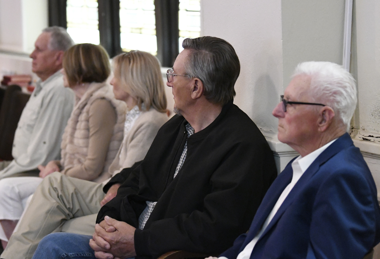 Several older adults sit in a row indoors, attentively listening to something off-camera, with natural light coming through windows behind them.