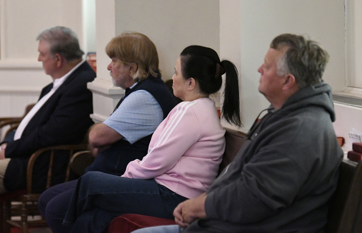 Four adults sit in a row, attentively listening indoors, with neutral expressions.