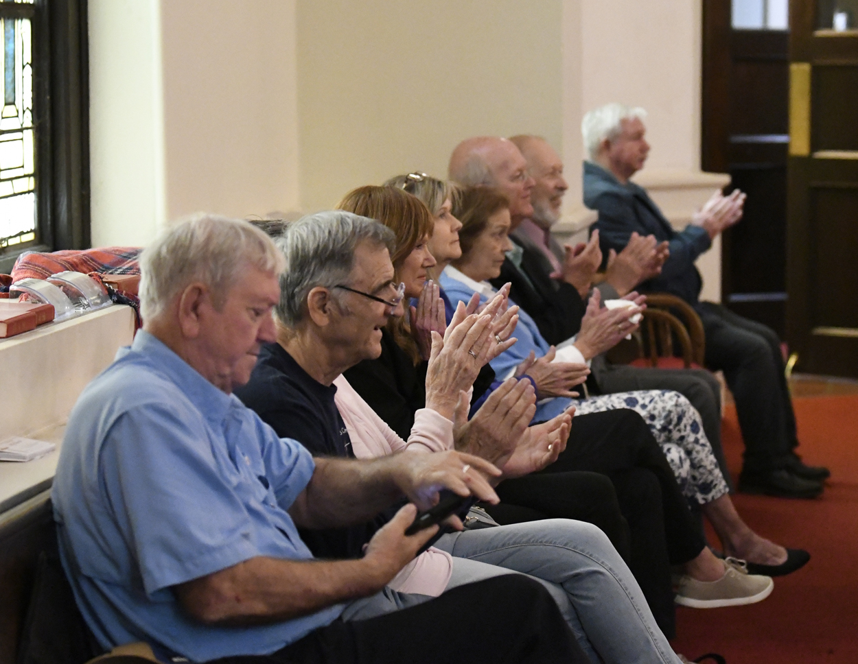 A group of elderly people sitting in rows indoors, clapping and smiling, appearing to attend an event or gathering.