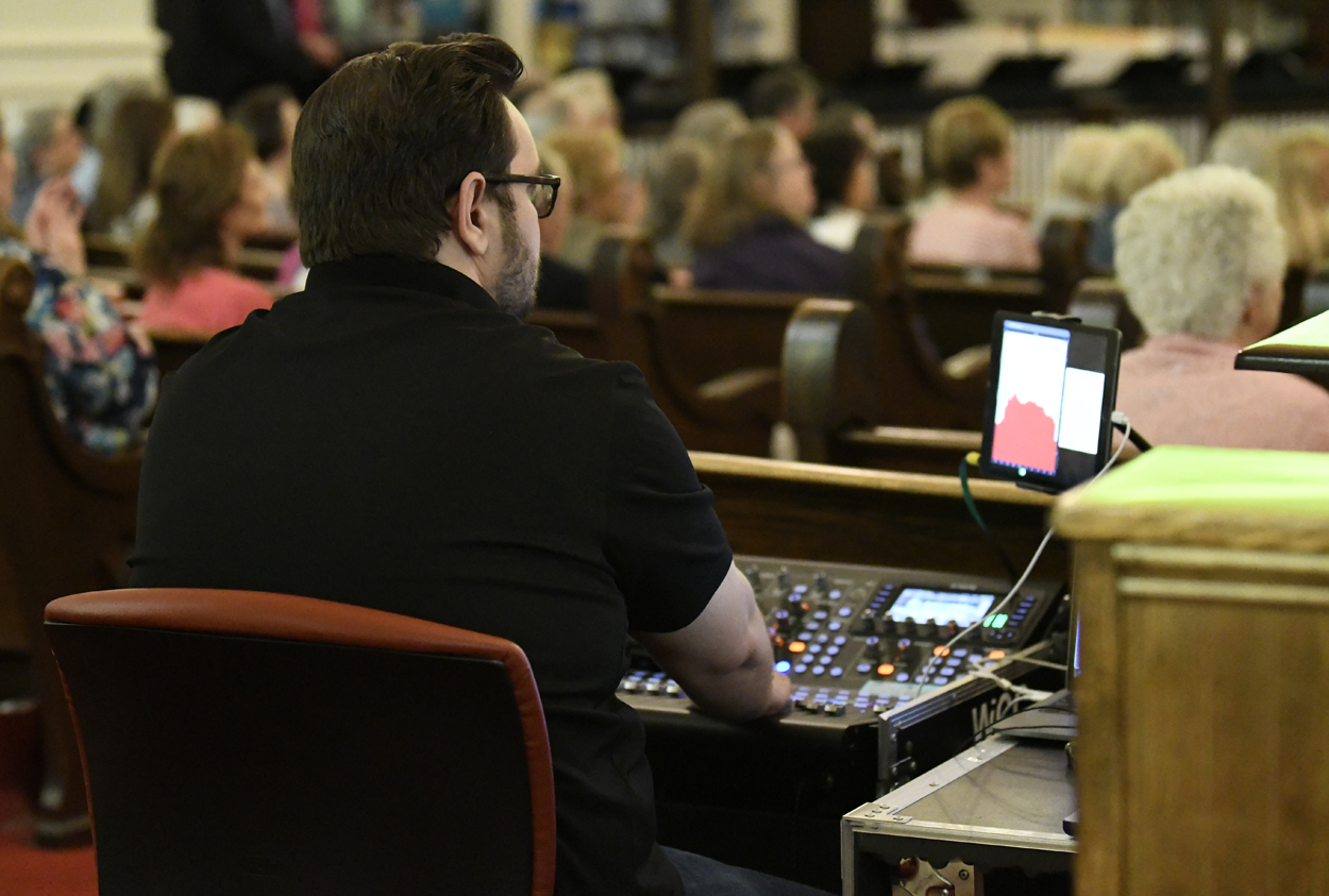 A man operating a sound mixing console at an indoor event, with an audience seated in the background.