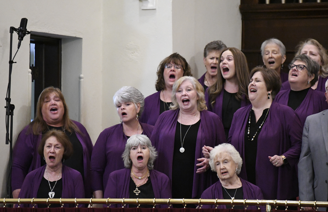 A group of women in matching purple outfits sing together in a choir, standing indoors with focused and expressive faces.