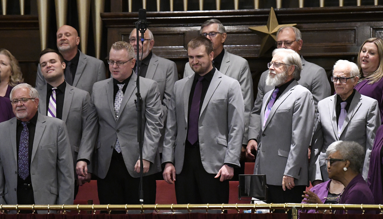 A group of men in matching gray blazers with black shirts and ties stand together singing in a choir setting.