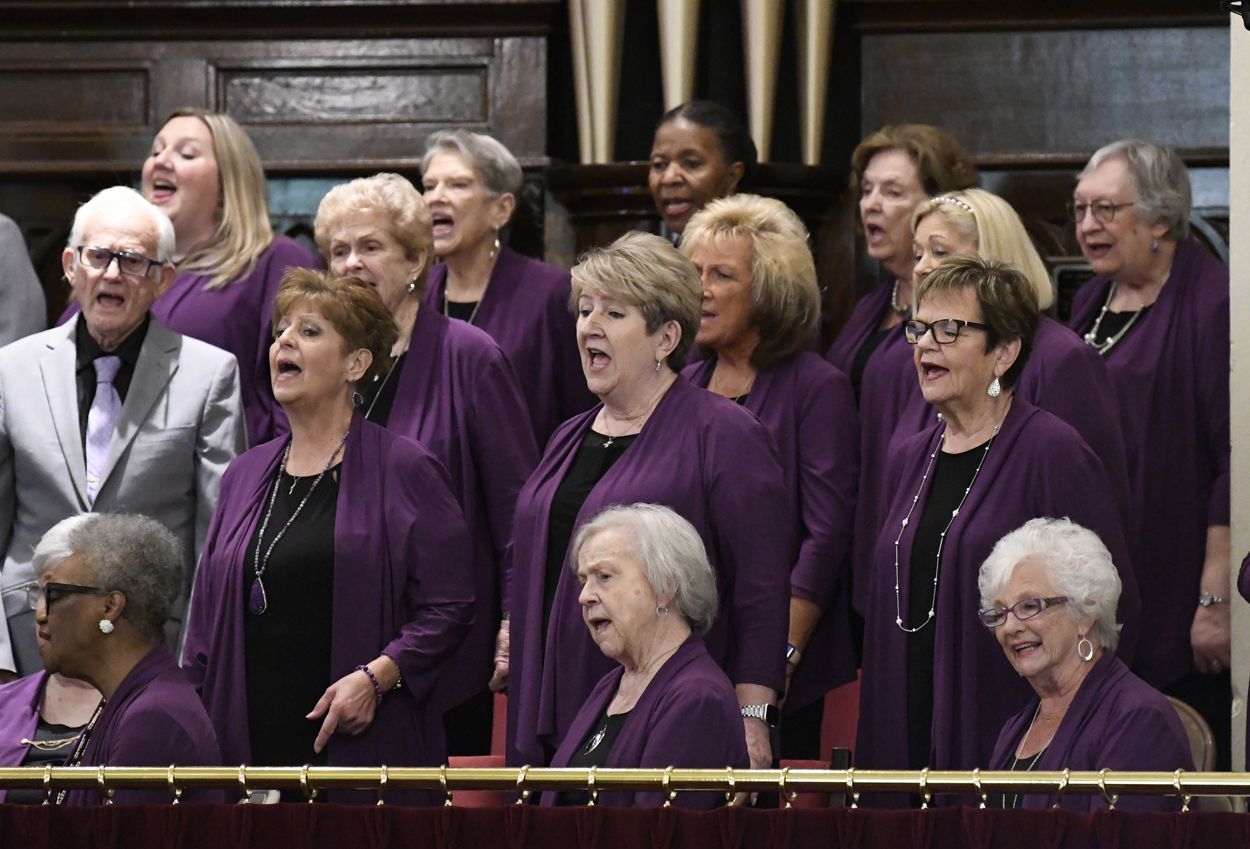 A group of women choir singers stand together, wearing matching purple outfits, performing in what appears to be a church or concert hall setting.