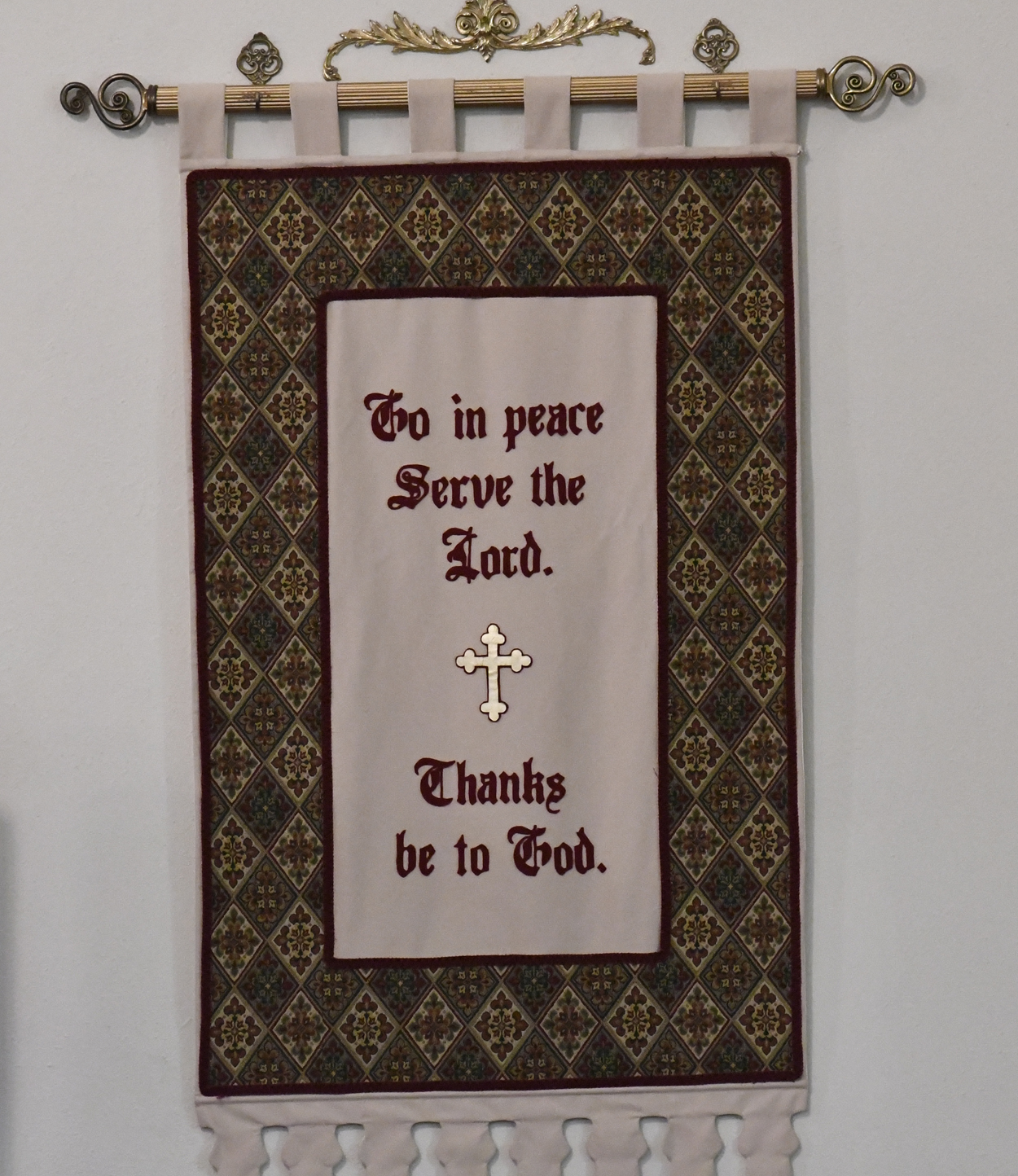 A decorative wall hanging with a cross and the words "Go in peace. Serve the Lord. Thanks be to God." embroidered in red text.