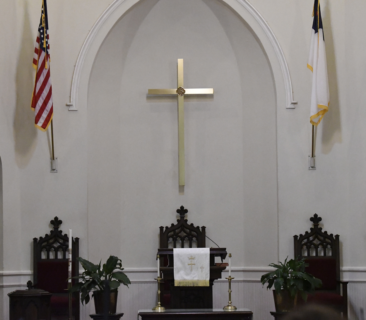 Church altar with a gold cross on the wall, flanked by American and Christian flags, and three ornate chairs below.