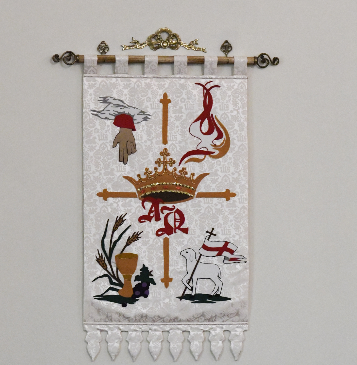 Embroidered wall hanging featuring a crown, cross, lamb with flag, hand, wheat, and decorative red motifs on a white background, finished with tassels at the bottom.