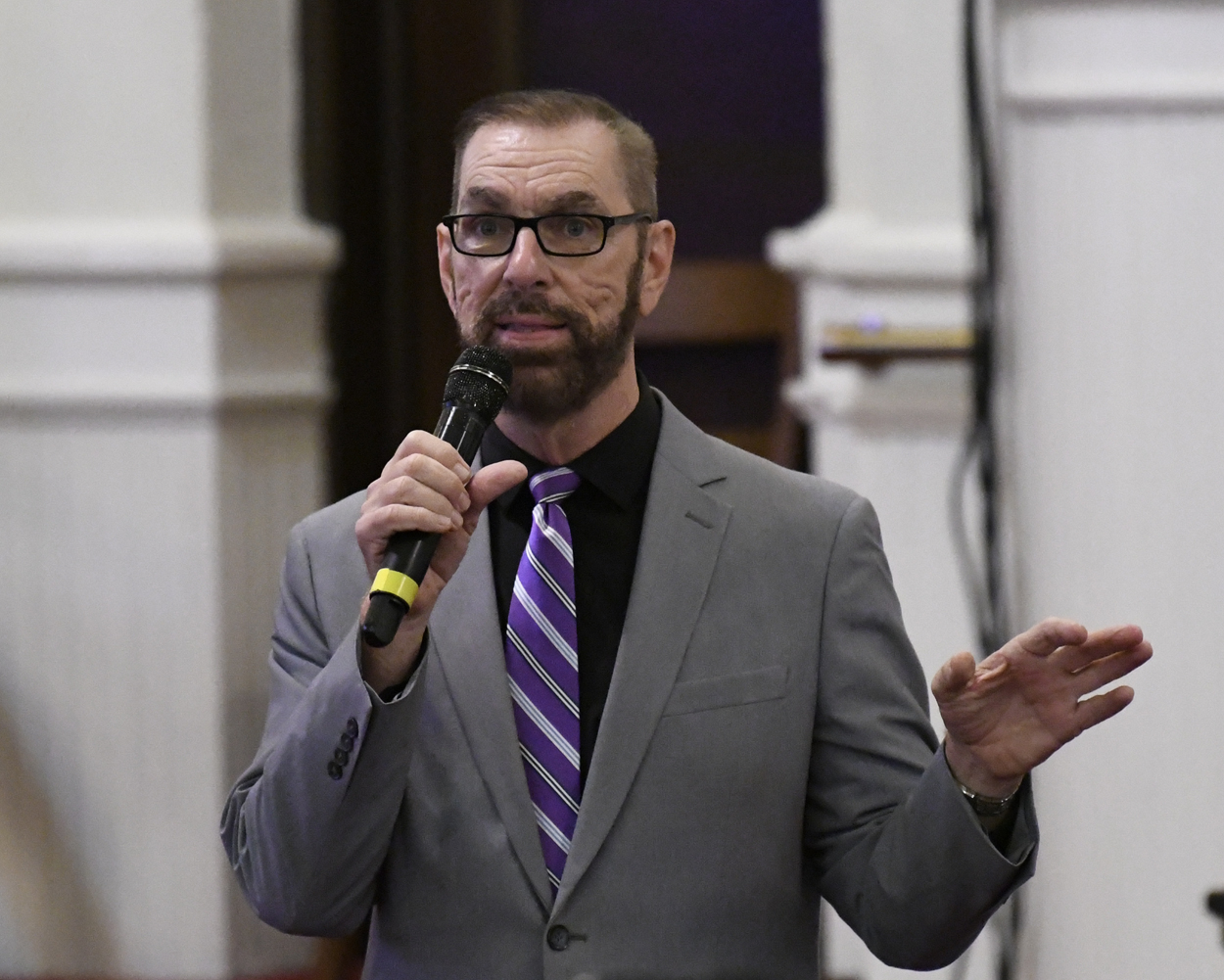 A man with glasses and a beard speaks into a microphone, wearing a gray suit and purple striped tie, standing indoors.