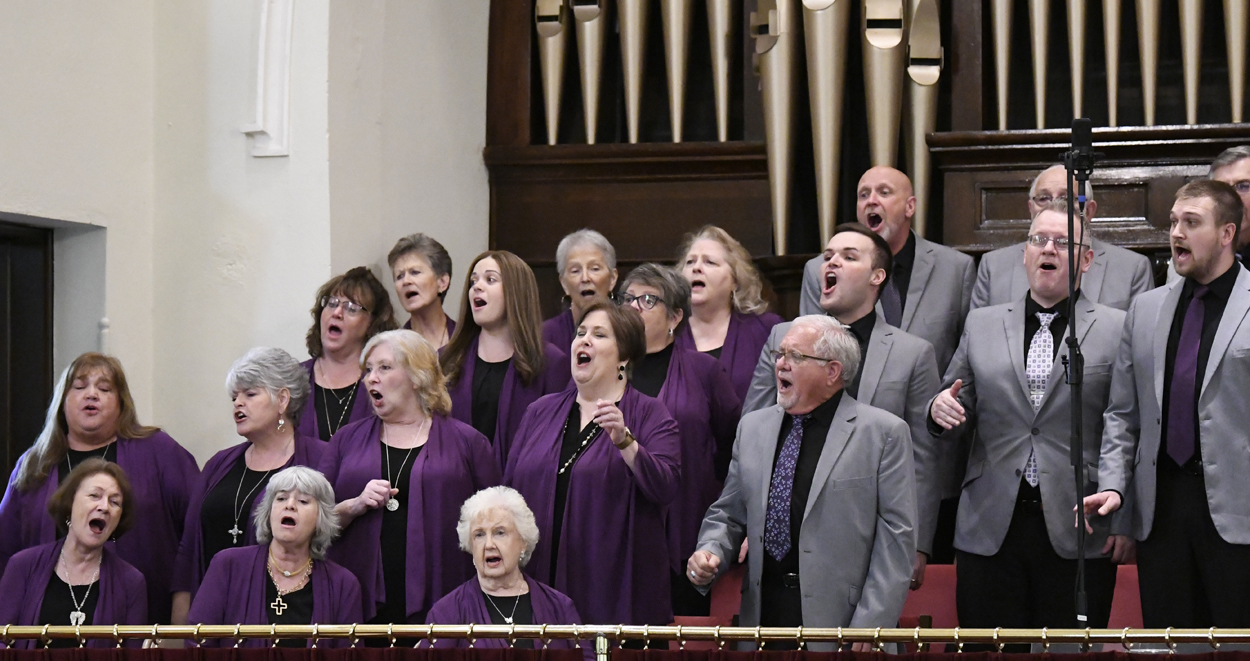A choir of men and women singing passionately in church, wearing purple robes and gray jackets, standing in front of large organ pipes.