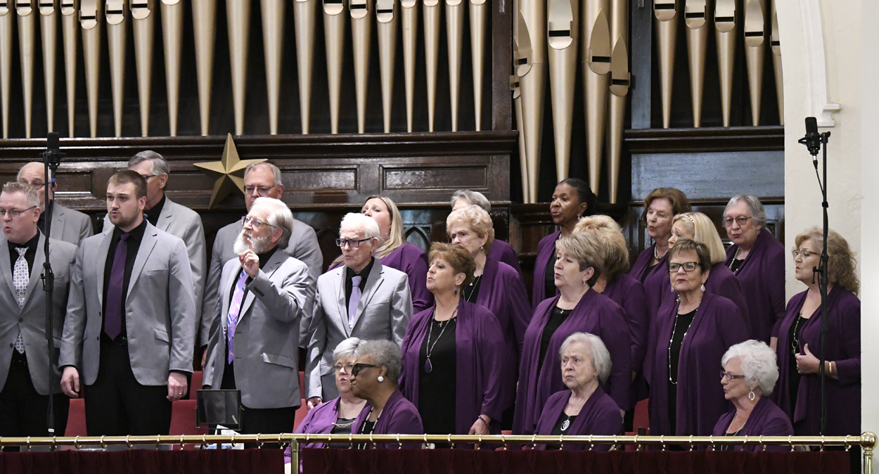 A choir of men and women, mostly dressed in purple, stands in front of a large pipe organ, singing during a performance inside a church.