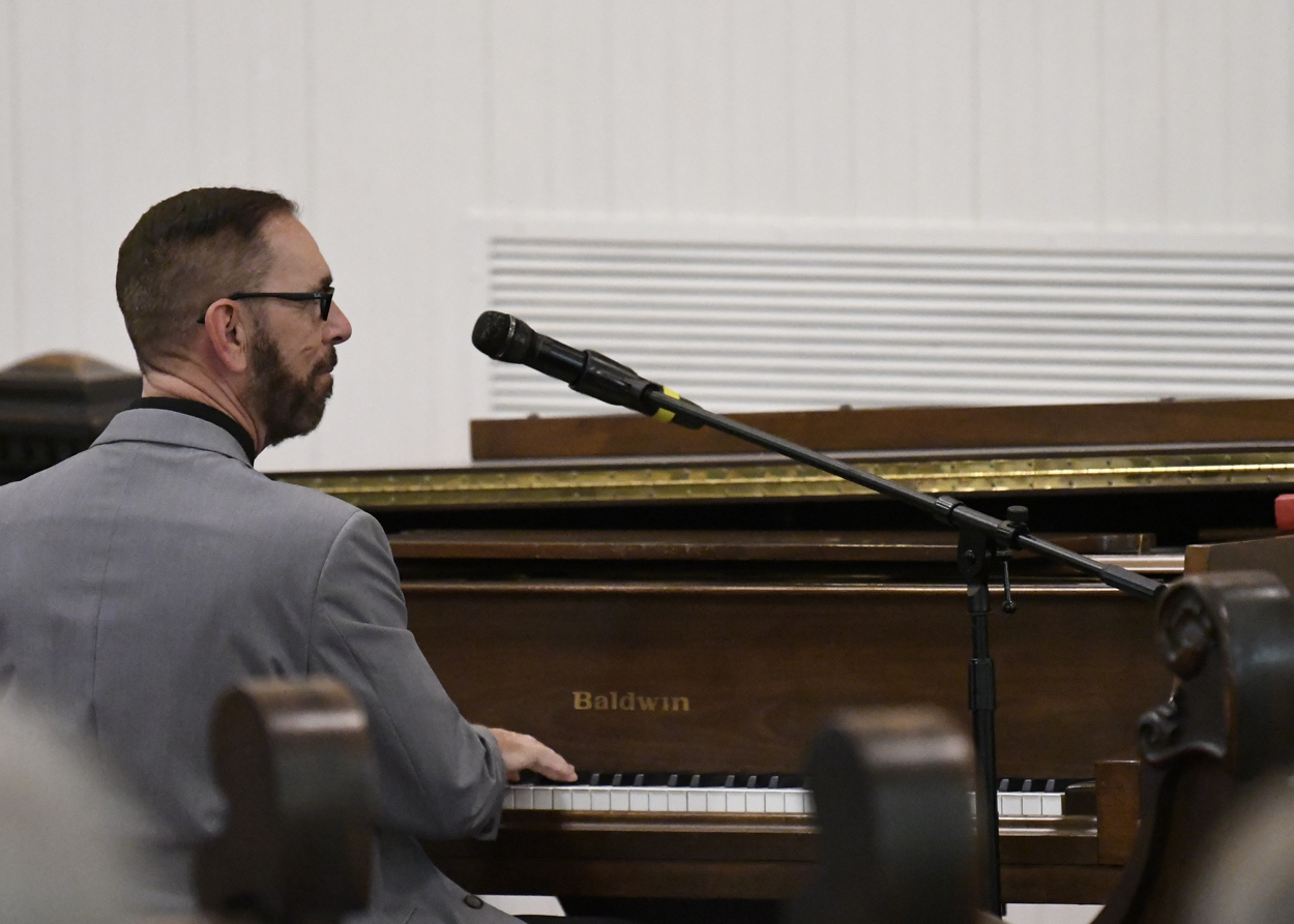 A man in a suit playing an upright piano and singing into a microphone in a church or hall setting.